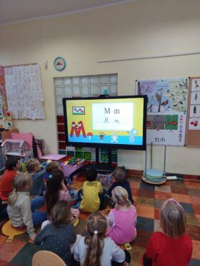 Dzieci siedzą przed ekranem. Na Monitorze włączona jest bajka edukacyjna o Myszce Mariannie, która prezentuje, jak piszemy małą i wielką literę M
