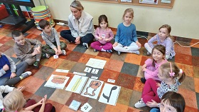 Dzieci siedzą z Panią w kole. Na środku rozłożone są obrazki przedstawiające nutę, torbę, termometr i tort oraz graficzny zapis litery T. 