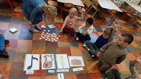 Dzieci siedzą w kole, w którym rozłożone są obrazki na literę T. Obok rozsypane są wszystkie litery alfabetu. Dziewczynka w niebieskiej sukience szuka pośród nich litery T.