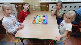 Cztery dziewczynki siedzą przy stoliku i uśmiechają się do zdjęcia. Na środku znajduje się stworzone przez nie mata sensoryczna, która składa się z piór, pokrywek, pianek i gąbek.