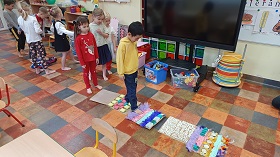 Na podłodze rozłożona jest stworzona przez dzieci mata sensoryczna. Chłopiec w żółtej bluzie idzie po macie, a reszta grupy czeka ustawiona w rzędzie. 