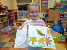 Uśmiechnięta dziewczynka trzyma przed sobą swoje prace. Pierwsza praca to kolorowy rysunek a druga to "ptaszek w gnieździe" wykonany za pomocą kolorowych liści. W tle widać bawiące się dzieci. 