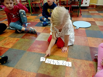 Na podłodze siedzi dziewczynka, która układa cyfry od 1 do 6. Wokół niej siedzą dzieci, które na nią spoglądają. 