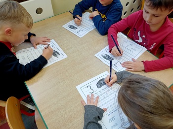 Przy stoliku siedzą dzieci, które piszą ołówkami cyfrę 3.
