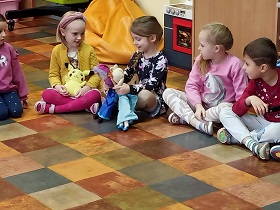 4 dziewczynki i 1 chłopiec siedzą na podłodze. Jedna dziewczynka prezentuje przyniesione przez siebie lalki. Pozostali ja obserwują. 