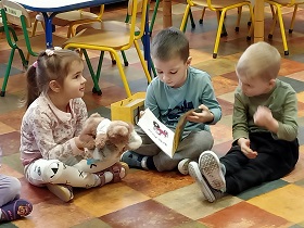 3 dzieci siedzi na podłodze - 1 dziewczynka i 2 chłopców. Dziewczynka trzyma przed sobą pluszowego misia. Jeden z chłopców trzyma przed sobą otwartą książkę. 