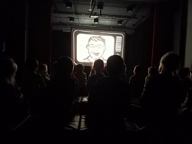 W ciemnej sali siedzą dzieci, które oglądają teatr cieni. Na białym ekranie znajduję się postać pana w monitorze.