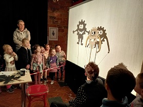 Dzieci stoją w wyznaczonej czerwoną taśmą przestrzeni i obserwują ekran, na którym pan w okularach pokazuje im szablony postaci, używane do teatru cieni.