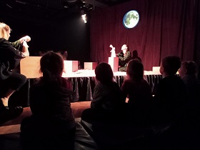 Dzieci siedzą na kolorowych poduszkach w przyciemnionej sali i obserwują to, co dzieje się na scenie, na której występują dwie aktorki ubrane na czarno i trzymają w dłoniach drewniane figurki.