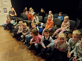 Dzieci siedzą na poduszkach i krzesełkach wraz z dorosłymi, w sali teatralnej. 