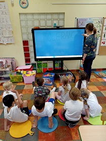 Dzieci siedzą na kolorowych poduszkach i spoglądają na monitor, na którym pani w bluzie w kwiatki pisze napis tolerancja. Napis ten znajduje się na niebieskim tle. 