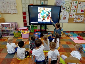 Dzieci siedzą na poduszkach przed monitorem, na którym wyświetlane jest zdjęcie dzieci, które trzymają się za ręce i otaczają świat.