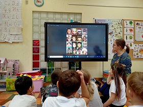 Dzieci siedzą przed monitorem i spoglądają na zdjęcie, na którym widać dzieci o różnych kolorach skóry i wyglądzie. 
