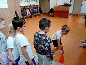 Dzieci w strojach sportowych stoją jedno za drugim. Jeden chłopiec trzyma rękę na pomarańczowym pachołku. 