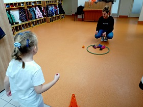 Dziewczynka w stroju sportowym stoi przy pomarańczowym pachołku. Przed nią przy hula hop kuca pani w czarnej bluzie. W hula hop leżą kolorowe woreczki. 