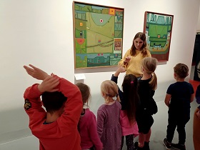 Dzieci wraz z panią w żółtym swetrze stoją przed obrazem, na którym znajdują się zielone kwadraty mniejsze i większe. Chłopiec w czerwonym swetrze się zgłasza.