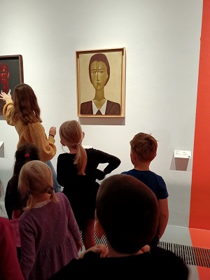 Dzieci wraz z panią w żółtym swetrze stoją przed obrazem, na którym znajduje się czyjś portret. Pani wskazuje na drugi obraz, na którym jest postać w czerwieni.