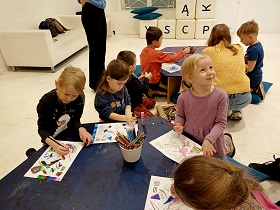 Dzieci siedzą przy stolikach i malują obrazki. Na stolikach stoją kredki w kubku oraz leżą farby w postaci pisaków. 
