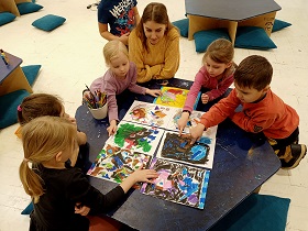 Dzieci wraz z panią w żółtym swetrze siedzą przy stoliku i oglądają prace plastyczne, które sami wykonali. 