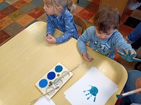 Dwie dziewczynki siedzą przy stole. Na stoliku leży paletka z 3 odcieniami niebieskiej farby, pędzelki oraz biała kartka z odbitą niebieską dłonią. Jedna z dziewczynek trzyma dłoń pomalowaną na niebiesko w górze. 