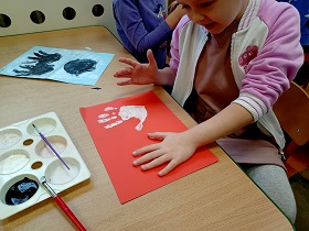 Dziewczynka odciska swoje dłonie pomalowane na biało na czerwonej kartce. Na środku stołu stoi paleta z farbami. A obok niej leży kartka z namalowanym farbami duchem.