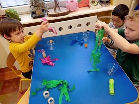 Troje chłopców siedzi przy stolikach i łączy ze sobą fragmenty różnych kolorów bibuły. Obok nich znajdują się plastikowe kubki. Na stoliku leżą też dwie taśmy klejące oraz kleje.