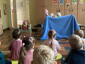 Dzieci siedzą na podłodze, przed nimi widać niebieski koc rozwieszony między dwoma krzesłami. Na kocu ułożona jest lalka i żabka. Z lewej strony pojawia się różowa pacynka zajączek.