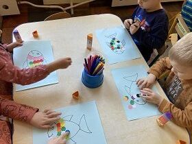 Czworo dzieci siedzi przy stoliku i przykleja kolorowe kółka wypełniając tułów ryby.