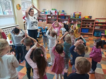 Dzieci tańczą w sali. Przed nimi stoi nauczycielka. Wszyscy mają uniesione, połączone nad głową ręce. 