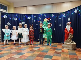 Na scenie stoją cztery dziewczynki przebrane za aniołki, dziewczynka przebrana za renifera, dwóch chłopców przebranych za elfy i chłopiec przebrany za Świętego Mikołaja.