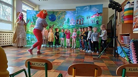 4 Dzieci stoją przed dekoracją obrazującą Krainę grzeczności w rzędzie. Z ich lewej strony stoi wróżka w złotej sukience i różowym kapeluszu. Postać z teatrzyku Zuzia próbuje złapać piłkę rzuconą przez dzieci. Ma pomarańczowe włosy, bluzkę w paski i czerwone spodnie.