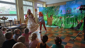 Wróżka w złotej sukience i różowym kapeluszu nachyla się do dzieci. Zuzia, która ma pomarańczowe włosy, bluzkę w paski i czerwone spodnie wyciera ręce w suknie wróżki.