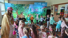Dzieci wraz z dwiema aktorkami tańczą w kole. Za nimi stoi dekoracja obrazująca Krainę Grzeczności. Dzieci kłaniają się do siebie. 