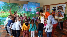 Dzieci wraz z dwiema aktorkami tańczą w kole. Za nimi stoi dekoracja obrazująca Krainę Grzeczności. Dzieci kłaniają się do siebie. 