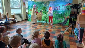 Z lewej strony stoi kobieta ubrana w złotą sukienkę i różowy kapelusz, która jest wróżką. Obok niej stoi kobieta w pomarańczowej peruce ubrana w bluzkę w paski i czerwone spodnie i gra na flecie prostym.