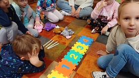 Dzieci siedzą w kole. Na środku leżą 4 maskotki, 4 ołówki i ułożone puzzle piankowe z oznaczeniami o zera do czterech.