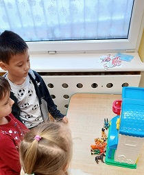 Dwóch chłopców i dziewczynka stoją przy stoliku, na którym stoi domek dla lalek. Obok domku ułożonych jest 6 figurek.