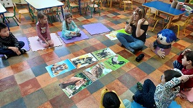 Dzieci siedzą na kolorowych matach w kole. Na środku rozłożone są obrazki słonia, koguta, kota, sowy, psa i konia.