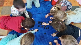 Dzieci siedzą na niebieskim miękkim kocu. Dotykają czarne i śliskie kamienie.