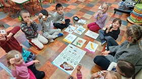 Dzieci siedzą w kolei pokazują odnalezione litery R. Na środku koła ułożone są obrazki robota, rakiety, róży, roweru, rajstop i plakat edukacyjny z literą R.