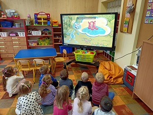 Dzieci siedzą w sali na podłodze. Przed nimi stoi tablica multimedialna z wyświetloną bajką o wodzie. 