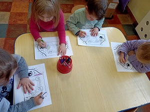 Cztery dziewczynki siedzą przy stoliku i kolorują rysunek przedstawiający kroplę wody oraz obieg wody w przyrodzie. Na środku stołu stoi kubek z kredkami. 