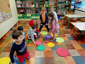 Dzieci stoją na środku sali i segregują kółka o różnych kolorach, układając je na odpowiednie kupki. 