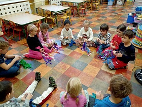 Dzieci siedzą na podłodze i trzymają w dłoniach plastikowe kubki z przyklejoną gumką recepturką i kolorową bibułą. Dzieci na nich grają.