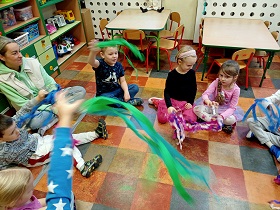 Dzieci unoszą swoje kubki plastikowe z kolorową bibułą i nimi ruszają. Obok dzieci siedzi też pani w zielono-białym stroju. 