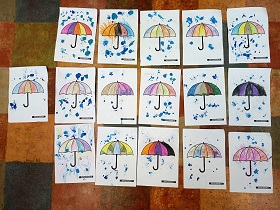 Na podłodze leży 16 rysunków przedstawiających pokolorowane parasole i padający deszcz namalowany za pomocą rozdmuchiwanej niebieskiej farby. 