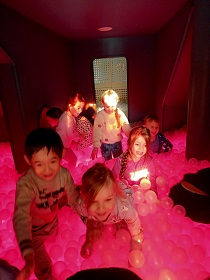 Dzieci znajdują się w podświetlanym na różowo basenie z białymi kulkami. 