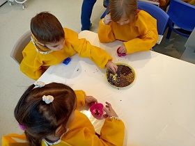 Dzieci ubrane w żółte fartuszki siedzą przy stoliku. Na środku stoi talerz z ziarnami kawy, różnymi kwiatkami i sypkimi materiałami. Każde dziecko przed sobą ma kolorową silikonową foremkę. Dzieci wypełniają foremki sypkimi materiałami. 