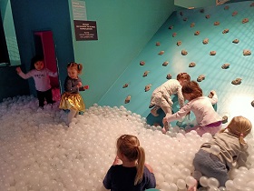 6 dziewczynek bawi się w strefie z plastikowymi kulkami. 2 z nich próbuje się wspinać po ściance wspinaczkowej. 