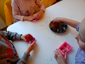 Troje dzieci siedzi przy stole i nakłada nasionka do silikonowych, różowych foremek. 